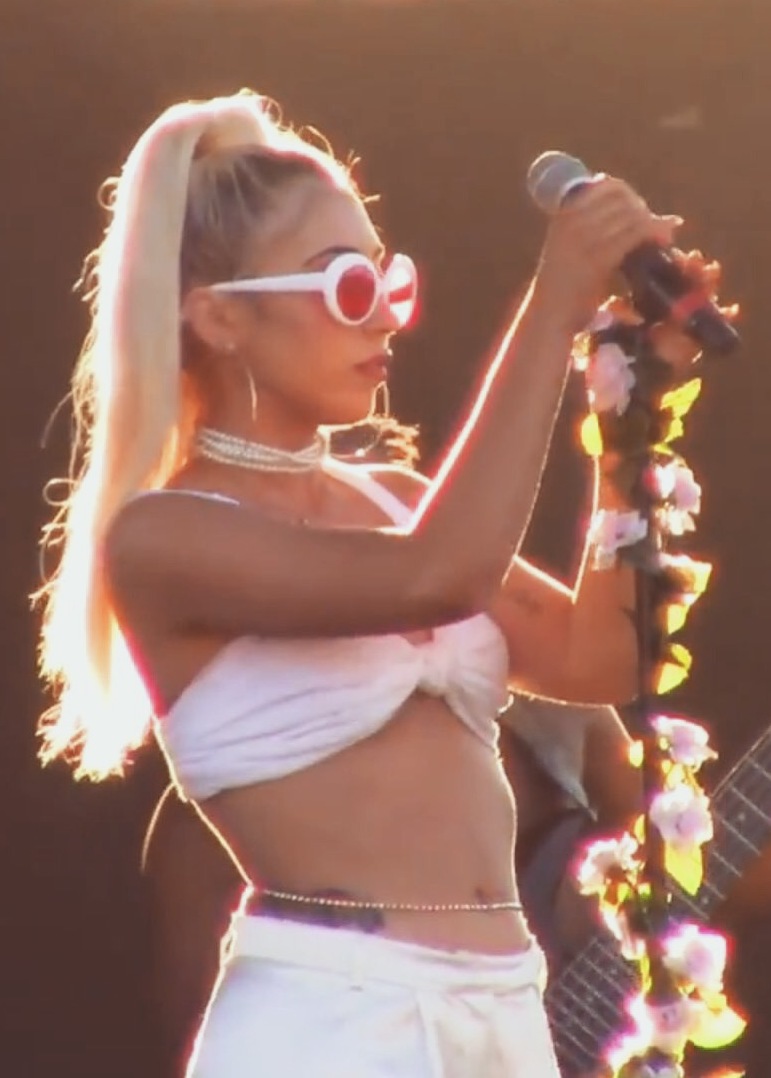 A singer wearing white sunglasses and a cropped top performs on stage, holding a microphone adorned with flowers, with sunlight creating a glow around her.